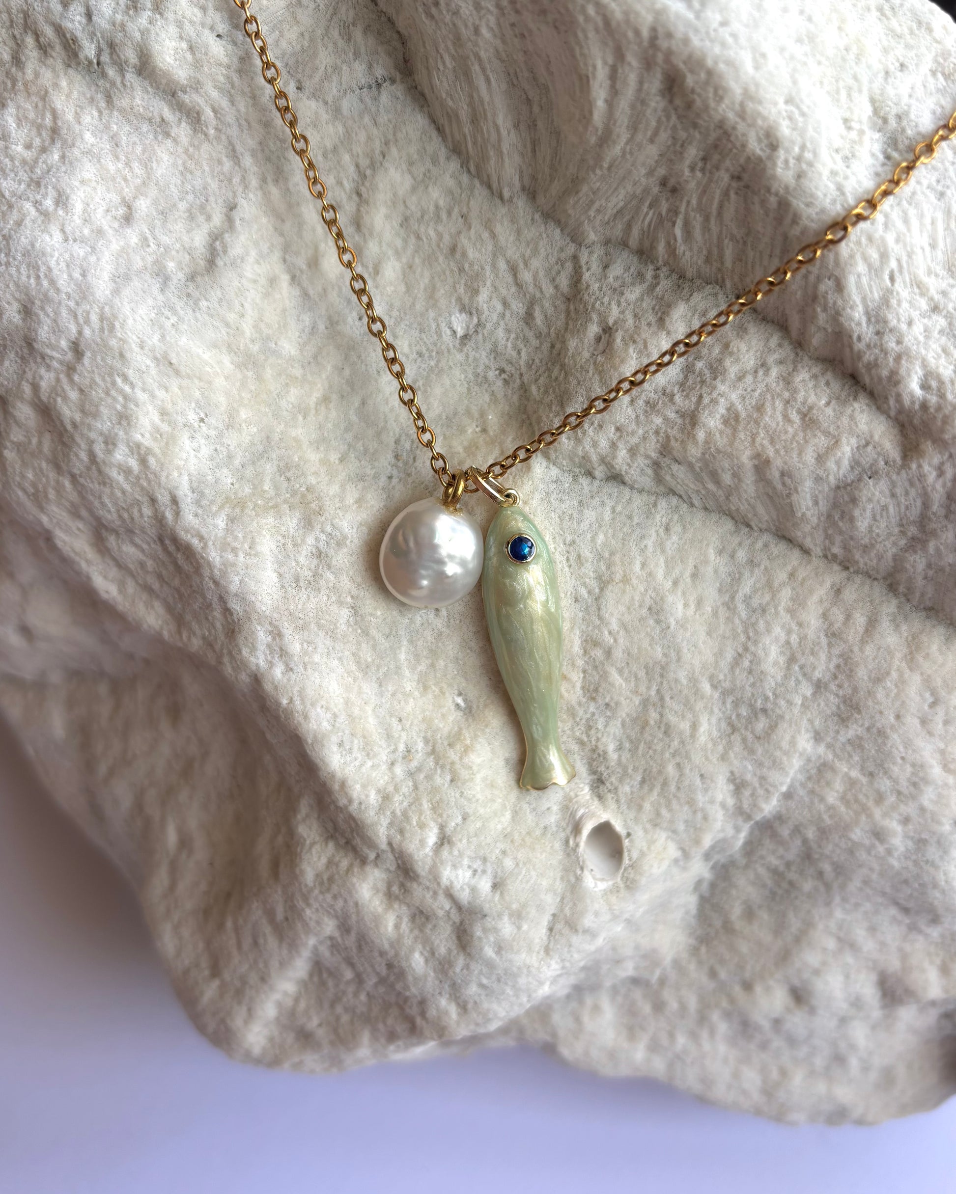Silhouette Spark CLOE necklace 近距離特寫，金色鍊條搭配白色陶瓷魚吊墜與藍眼珍珠圓珠，石材紋理背景